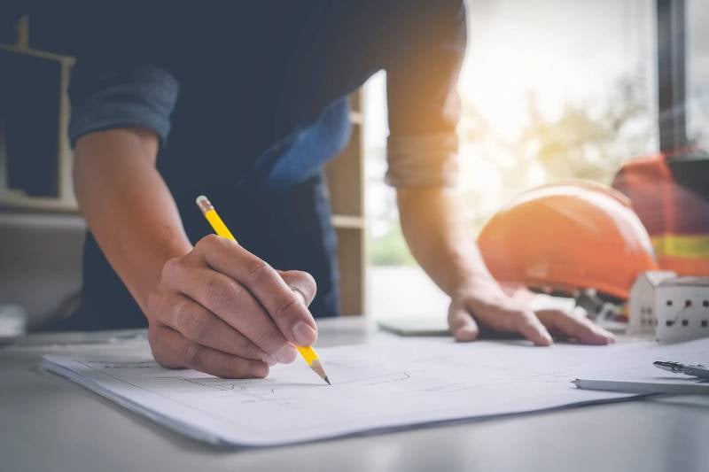 Person drawing architectural blueprints with a pencil, a hard hat and model house in the background.