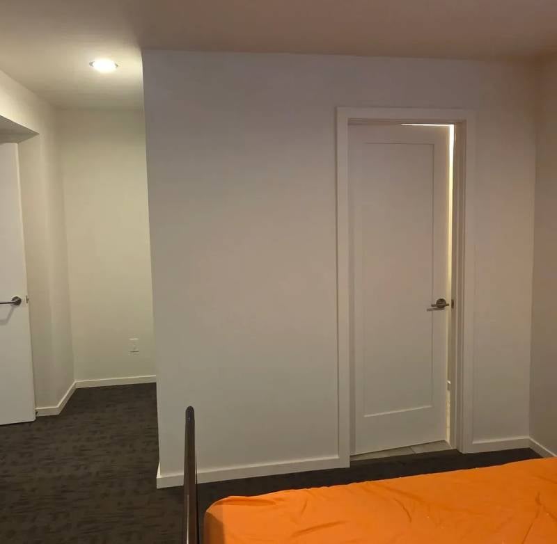 A bedroom with an orange bedspread, white walls, a partially open door with light shining through, and a hallway beyond.
