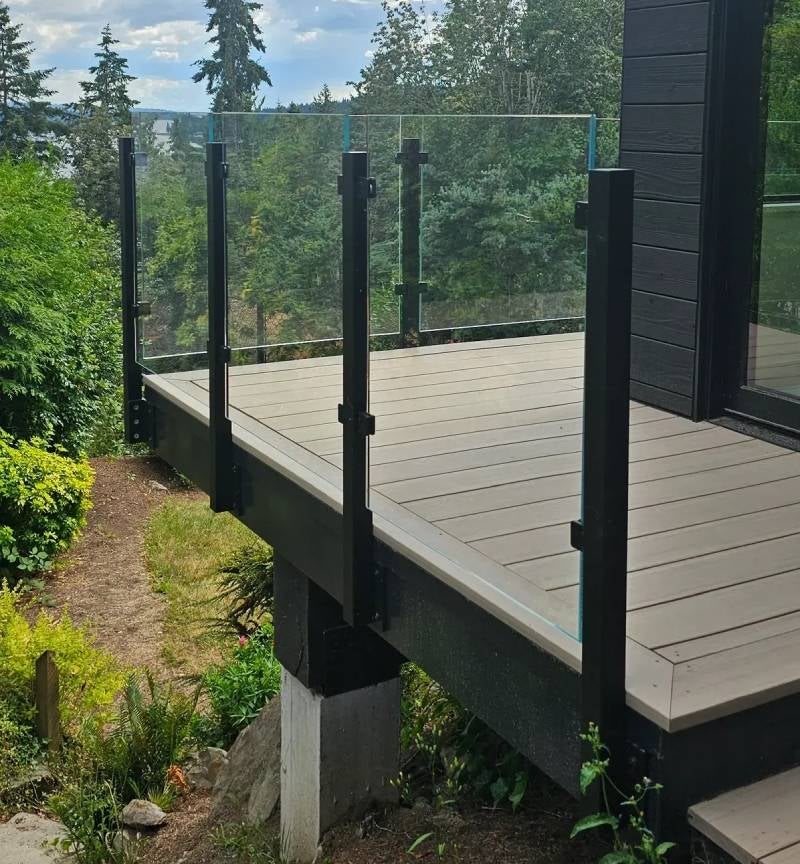 Modern deck with light brown boards, black-posted glass railing, overlooking a lush green forest and distant water.