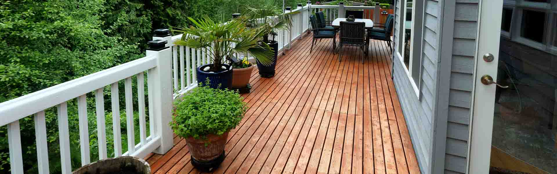 Wooden deck with white railings, potted plants, and outdoor furniture, overlooking lush green trees.